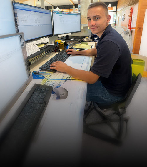 Stanglmair Auszubildender sitzt am Schreibtisch vor mehreren Monitoren und schaut in die Kamera